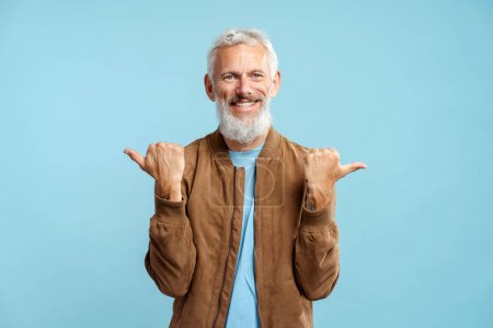 Foto de Retrato de hombre de pelo gris barbudo sonriente con elegante chaqueta marrón casual señalando los dedos mirando a la cámara, de pie aislado sobre fondo beige. Concepto de compras, publicidad - Imagen libre de derechos