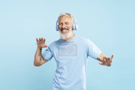Téléchargez les photos : Portrait d'un homme barbu mature souriant écoutant de la musique dans un casque aux yeux fermés dansant isolé sur fond bleu. Concept technologique, amusant - en image libre de droit