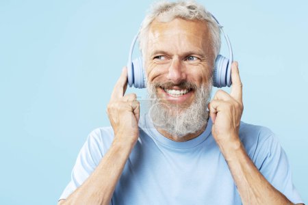 Téléchargez les photos : Portrait d'un bel homme mature aux cheveux gris, hipster écoutant de la musique dans des écouteurs regardant de loin gros plans isolés sur fond bleu, espace de copie. Concept technologique - en image libre de droit
