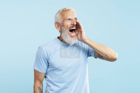 Photo for Portrait of mature angry bearded man shouting announcing something, looking away wearing stylish casual blue t shirt standing isolated on blue background. Communication concept - Royalty Free Image