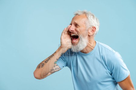 Photo for Portrait of mature bearded man shouting announcing something, looking away wearing stylish casual blue t shirt standing isolated on blue background copy space. Communication concept - Royalty Free Image