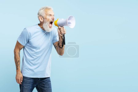 Foto de Enojado hombre de pelo gris maduro sosteniendo altavoz gritando algo, vistiendo camiseta azul casual de pie aislado sobre fondo azul. Concepto de protesta, comunicación - Imagen libre de derechos