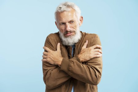 Foto de Retrato de un triste hombre maduro barbudo usando una elegante chaqueta marrón calentándose, mirando hacia otro lado parado aislado sobre un fondo azul. Concepto de tratamiento - Imagen libre de derechos
