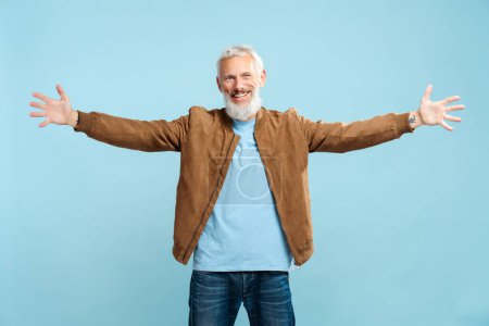 Foto de Retrato de hombre barbudo maduro sonriente con elegante chaqueta marrón casual abrazándose, mirando a la cámara, de pie aislado sobre fondo azul. Concepto de publicidad, gesto - Imagen libre de derechos