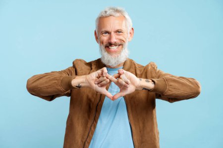 Foto de Retrato de sonriente atractivo barbudo hombre de pelo gris con elegante chaqueta marrón gesto con los dedos mirando a la cámara de pie aislado sobre fondo azul. Concepto de lenguaje corporal - Imagen libre de derechos