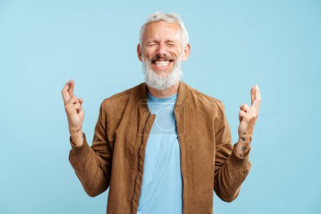 Téléchargez les photos : Portrait d'homme mature barbu aux cheveux gris souriants portant une veste décontractée élégante avec les doigts croisés et les yeux fermés debout isolés sur fond bleu. Concept d'espoir - en image libre de droit