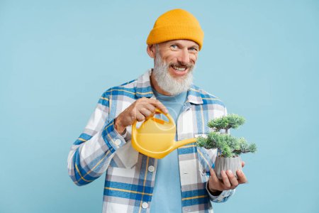 Foto de Retrato de un atractivo hombre de pelo gris con una camisa elegante, sosteniendo maceta de flores regándola con una regadera amarilla, mirando a la cámara de pie aislada sobre un fondo azul. Concepto de jardinería - Imagen libre de derechos
