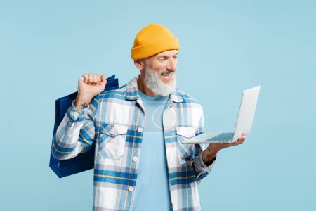 Foto de Retrato de un hombre maduro y atractivo que lleva una camisa elegante y una gorra amarilla, usando una computadora portátil y una bolsa de compras aisladas sobre un fondo azul. Pedidos en línea, concepto de compras - Imagen libre de derechos