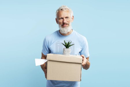 Photo for Upset mature bearded 50 year old man holding cardboard box with things,firing from job, looking at camera standing isolated on blue background. Concept of dismissal from work - Royalty Free Image