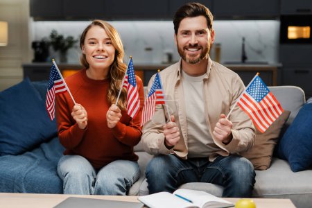 Téléchargez les photos : Un couple d'amis américains souriants tenant des drapeaux américains regardant la fête de l'indépendance assis à la maison. Liberté, 4 juillet - en image libre de droit