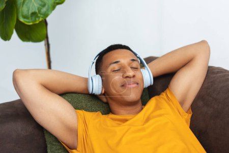 Foto de Retrato de un hombre afro-americano alegre y positivo con auriculares, escuchando música, tumbado en un cómodo sofá, relajándose con los ojos cerrados. Concepto de publicidad - Imagen libre de derechos