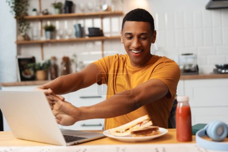 Téléchargez les photos : Portrait de beau jeune homme afro-américain souriant travaillant à la maison assis dans la cuisine, à l'aide d'un ordinateur portable, étirement, pause snack, sandwich savoureux. Travail à distance, concept alimentaire - en image libre de droit