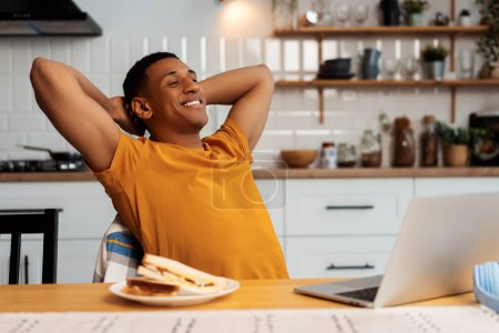 Téléchargez les photos : Portrait de jeune homme afro-américain souriant fatigué s'étirant tout en étant assis dans la cuisine moderne à la maison, en utilisant un ordinateur portable, une collation, un sandwich savoureux. Concept de déjeuner, pause - en image libre de droit