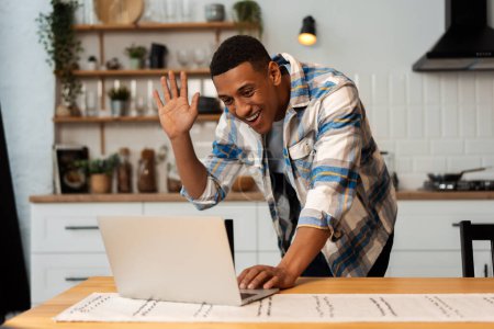 Téléchargez les photos : Attrayant souriant homme afro-américain, jeune homme d'affaires portant des vêtements décontractés travaillant à la maison, en utilisant un ordinateur portable, ayant appel vidéo agitant la main, debout dans un appartement élégant. Concept d'emploi à distance en ligne - en image libre de droit