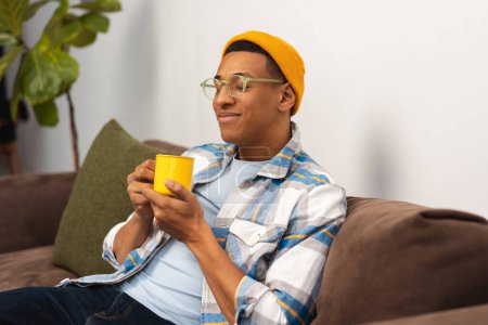 Téléchargez les photos : Portrait d'un homme afro-américain authentique portant un élégant chapeau jaune, tenant une tasse de café jaune, les yeux fermés assis sur un canapé confortable, se reposant. Concept relaxant - en image libre de droit