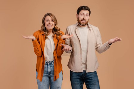 Photo for Clueless and puzzled stylish male and female standing at beige wall, having confused looks, shrugging shoulders as if saying: Who cares, I don't know, What's the problem - Royalty Free Image