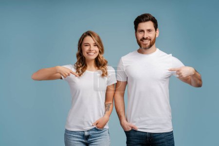 Téléchargez les photos : Portrait d'un couple souriant et confiant, hipsters vêtus d'un t-shirt blanc, pointant isolé sur fond bleu. Concept de maquette - en image libre de droit