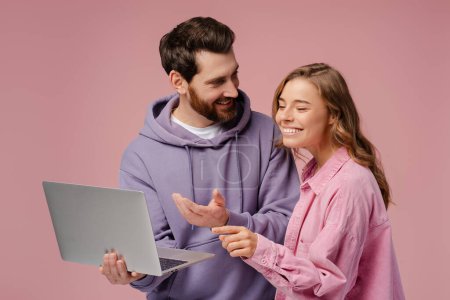 Foto de Un par de amigos sonrientes sosteniendo computadora portátil eligiendo bienes aislados sobre fondo rosa. Elegante hombre y mujer de compras en línea mirando a la pantalla. Concepto tecnológico - Imagen libre de derechos