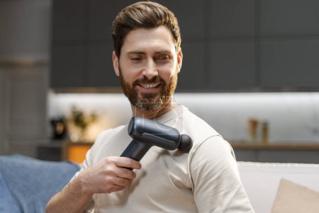 Photo for Portrait of middle aged bearded man using massage gun, doing self massage, massaging shoulder while sitting on comfortable sofa at home. Treatment concept, relaxing - Royalty Free Image