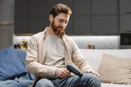 Photo for Portrait of attractive handsome bearded man using massage gun, doing self massage, massaging knee while sitting on comfortable sofa at home. Concept of health care, treatment, recovery - Royalty Free Image