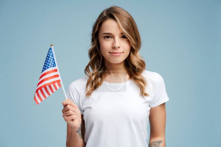 Téléchargez les photos : Portrait de mignonne, belle jeune femme, patriote, supporter avec coiffure élégante tenant drapeau américain portant des vêtements décontractés debout isolé sur fond bleu. Concept de jour de l'indépendance - en image libre de droit