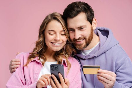 Téléchargez les photos : Portrait de couple souriant, attrayant homme et femme debout isolé sur fond rose, tenant le téléphone mobile, utilisant l'application mobile, tenant la carte de crédit, paiement. Concept d'achat en ligne - en image libre de droit