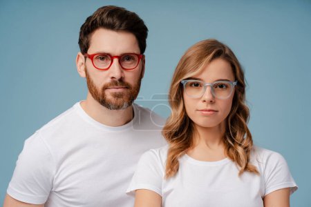 Photo for Closeup portrait of attractive serious man and woman wearing white t shirt and stylish eyeglasses isolated on blue background. Confident fashion models looking at camera posing for pictures in studio - Royalty Free Image