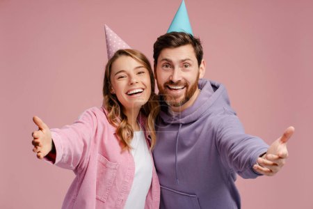 Téléchargez les photos : Couple d'amis heureux portant anniversaire cône célébration anniversaire isolé sur fond rose. Anniversaire, concept d'invitation de fête - en image libre de droit