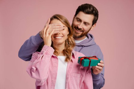 Photo for Handsome smiling man holding gift box, closed eyes of young beautiful woman. Portrait of happy romantic couple celebration birthday isolated on pink background. Dating, love, Valentines day - Royalty Free Image