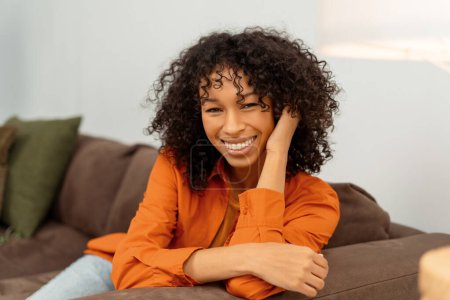 Téléchargez les photos : Portrait de belle femme afro-américaine souriante avec des cheveux bouclés élégants regardant la caméra assise sur un canapé confortable à la maison. Joyeux mannequin portant une chemise orange posant pour des photos - en image libre de droit