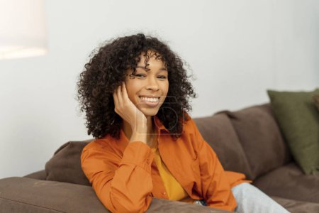 Téléchargez les photos : Authentique portrait heureuse belle afro-américaine aux cheveux bouclés relaxant à la maison, assise sur un canapé confortable. Concept de beauté naturelle - en image libre de droit