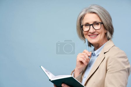 Foto de Mujer mayor sonriente y confiada en el retrato, maestra de escuela con anteojos con bolígrafo y bloc de notas aislados sobre fondo azul. Gerente de éxito feliz, CEO, empresaria mirando a la cámara - Imagen libre de derechos