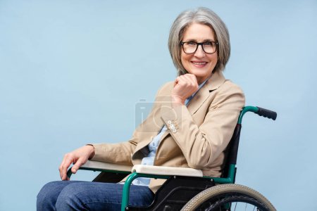 Foto de Retrato confiado sonriendo mujer mayor con gafas con estilo sentado en silla de ruedas mirando la cámara aislada sobre fondo azul - Imagen libre de derechos