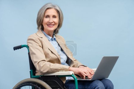 Foto de Retrato sonriente mujer mayor de pelo gris usando computadora portátil que trabaja en línea sentado en silla de ruedas aislado sobre fondo azul. Concepto de trabajo independiente - Imagen libre de derechos