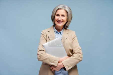 Téléchargez les photos : Portrait souriant femme âgée, gestionnaire, travailleur tenant ordinateur portable regardant la caméra isolée sur fond bleu - en image libre de droit