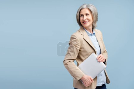 Foto de Retrato de mujer de negocios mayor sonriente con confianza, CEO con traje elegante que sostiene la computadora portátil mirando hacia otro lado aislado sobre fondo azul, espacio de copia. Concepto de negocio exitoso - Imagen libre de derechos