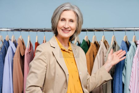 Photo for Smiling senior woman, stylist, fashion designer standing near clothes rail with a lot stylish clothes, showing wardrobe. Portrait successful tailor in atelier. Shopping, store, small business concept - Royalty Free Image