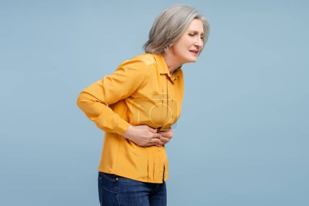 Téléchargez les photos : Femme âgée tenant ventre ayant mal au ventre isolé sur fond bleu. Diarrhée, douleur, gastrite, intoxication alimentaire concept - en image libre de droit