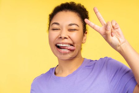 Téléchargez les photos : Portrait de femme afro-américaine souriante montrant la langue, geste de paix gestuel, s'amuser debout isolé sur fond jaune, gros plan. Concept de style de vie positif - en image libre de droit