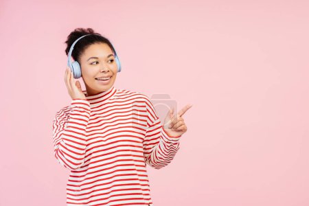 Foto de Retrato de una mujer afroamericana emocionada sonriente que usa un suéter elegante escuchando música en auriculares que señalan con el dedo el espacio de copia de pie aislado sobre un fondo rosa. Concepto de publicidad - Imagen libre de derechos
