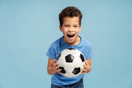 Foto de Retrato de niño guapo sonriente, jugador de fútbol con camiseta azul sosteniendo pelota de fútbol mirando a la cámara de pie aislado sobre fondo azul. Jugar al fútbol concepto - Imagen libre de derechos