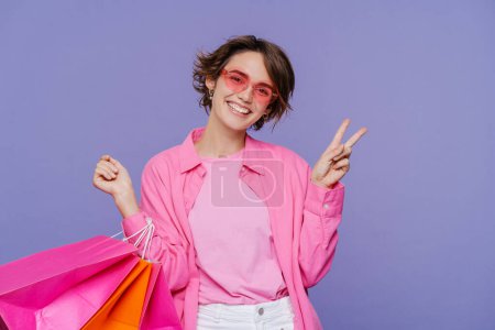 Foto de Joven mujer feliz con el pelo corto en ropa rosa sosteniendo coloridas bolsas de compras aisladas sobre fondo violeta. Tienda, compras, ventas, concepto cyber Monday - Imagen libre de derechos
