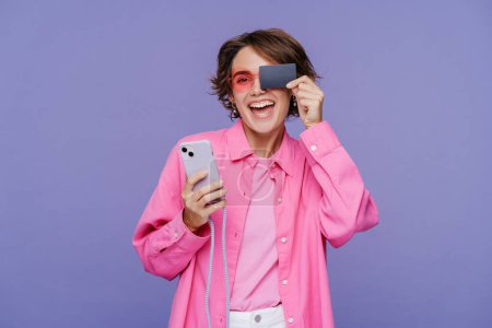 Photo for Smiling modern woman holding mobile phone and covering eye with credit card, online shopping, isolated on violet background. Concept of mobile banking - Royalty Free Image