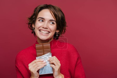 Foto de Joven mujer feliz en suéter rojo sosteniendo barra de chocolate y mirando hacia otro lado aislado en retrato de estudio de fondo rojo. Concepto de comida estilo de vida - Imagen libre de derechos