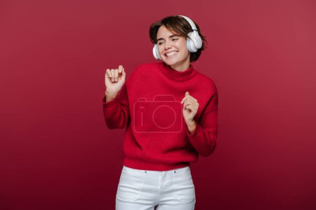 Téléchargez les photos : Belle femme souriante portant des écouteurs écoutant de la musique, regardant loin isolé sur fond rouge, dansant, s'amusant. Concept de mode de vie heureux - en image libre de droit