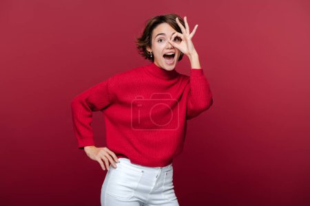 Foto de Retrato de mujer atractiva y elegante con elegante traje de suéter rojo que muestra signo de ok, mirando a la cámara con la boca abierta aislada en el fondo rojo - Imagen libre de derechos