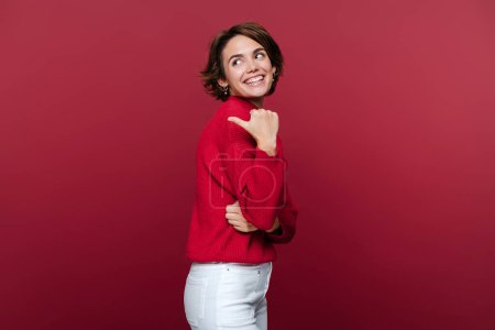 Téléchargez les photos : Portrait studio de belle femme souriante portant un pull rouge pointant de côté l'espace de copie pour votre texte publicitaire, sur fond rouge isolé - en image libre de droit