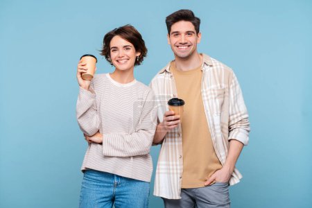 Téléchargez les photos : Portrait d'un beau couple souriant portant des vêtements décontractés tenant des tasses de café regardant la caméra boire isolé sur fond bleu. Concept de pause café - en image libre de droit