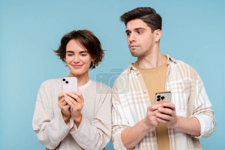 Téléchargez les photos : Homme intéressé regardant smartphone de sa petite amie isolée sur fond bleu. Amis heureux tenant des téléphones mobiles, communication en ligne. Technologie, concept de dépendance aux médias sociaux - en image libre de droit