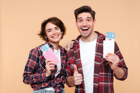 Photo for Cheerful couple passenger tourists going for weekend getaway, smiling at camera, posing over beige background with a passports and airplane tickets, awaiting to board the flight - Royalty Free Image